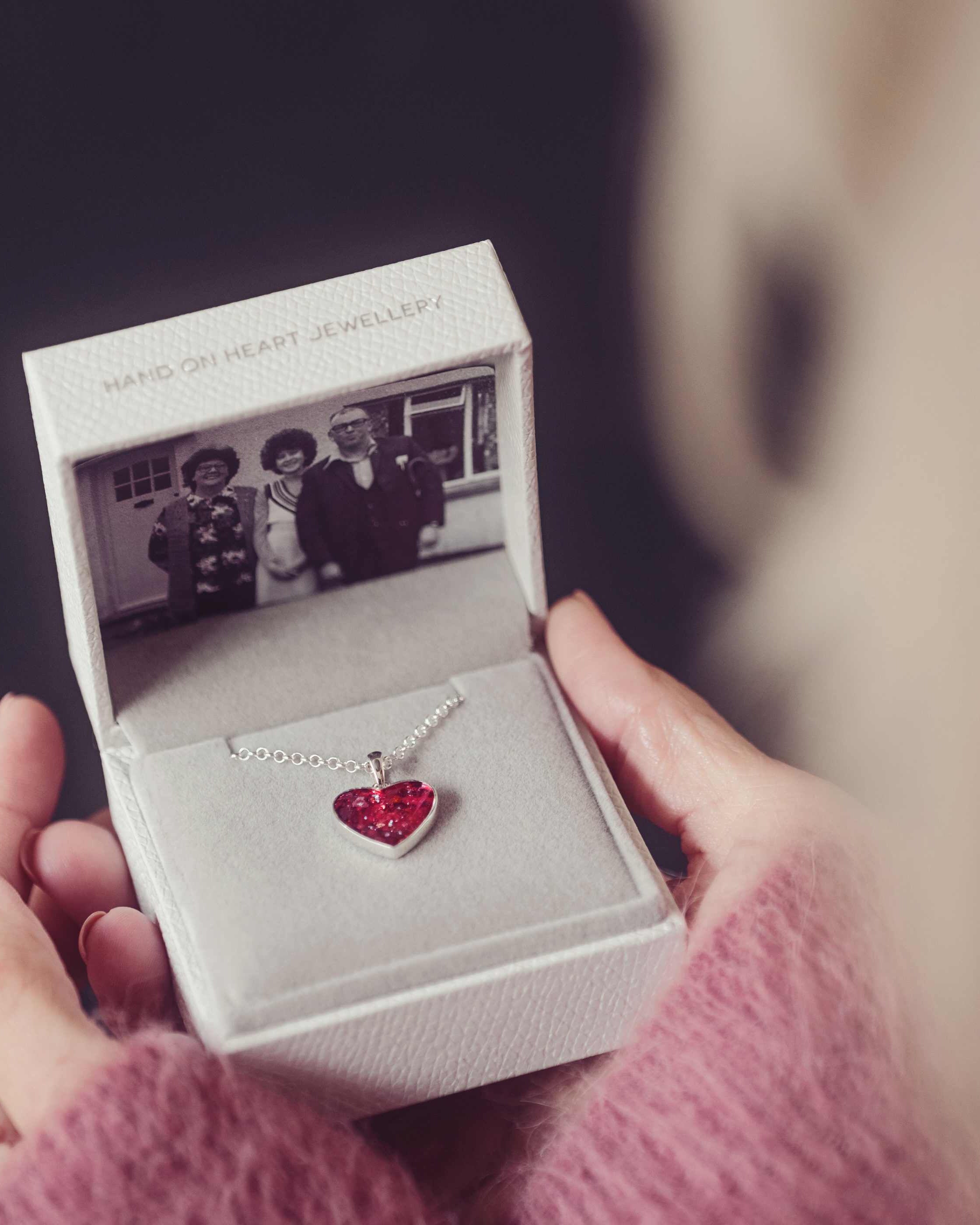 A red heart cremation ashes necklace being held in a grey jewellery box with a photograph of three people.