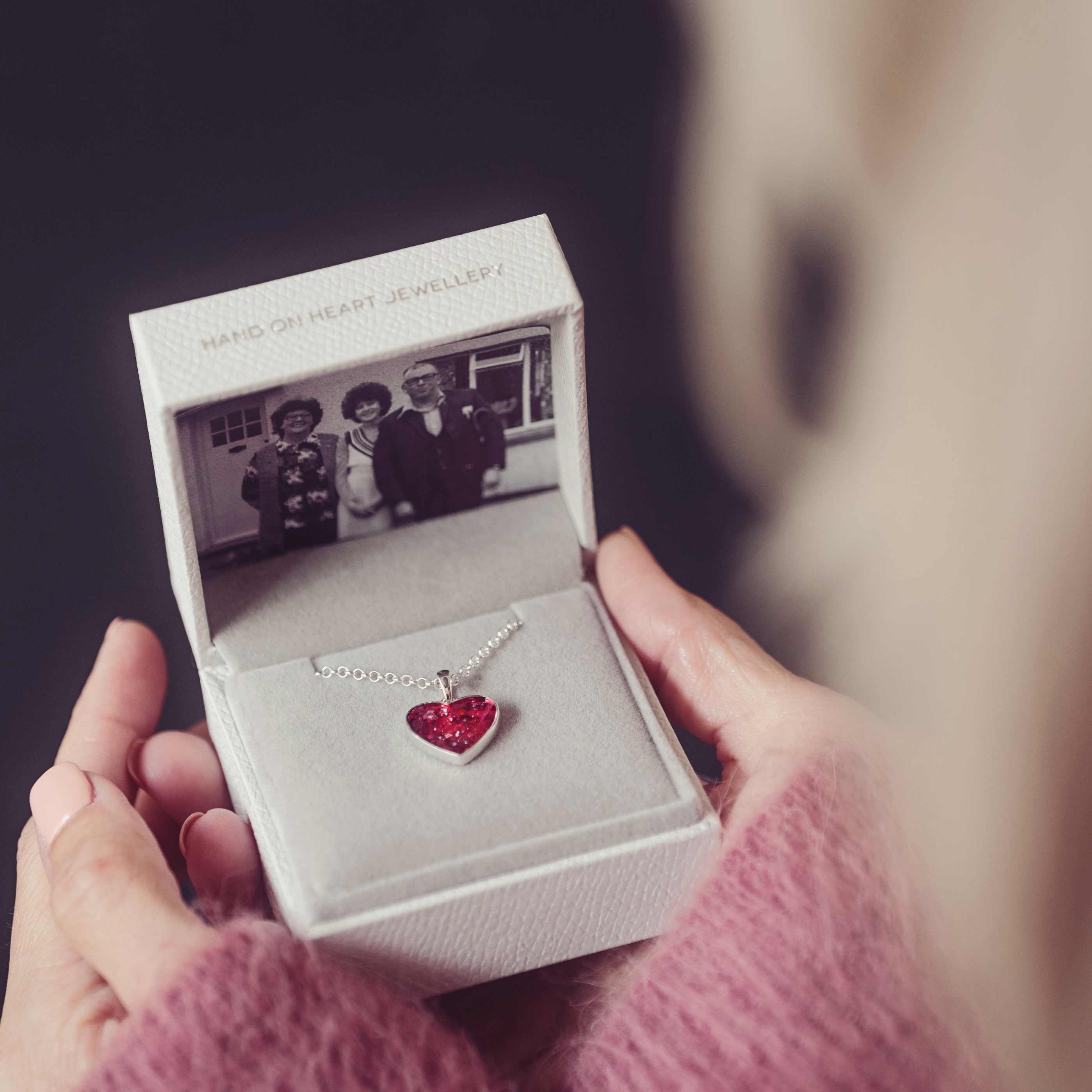 A red heart cremation ashes necklace being held in a grey jewellery box with a photograph of three people.