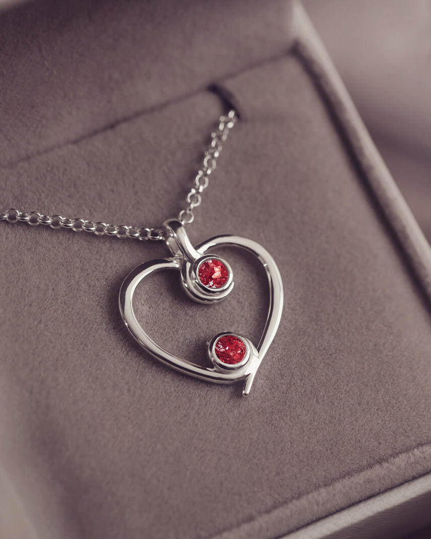 A silver heart-shaped pendant necklace featuring two sparkling red resin stones, holding cremation ashes, displayed against a neutral background.