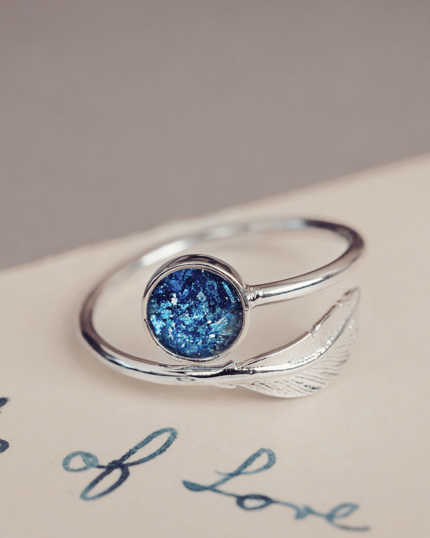 The image shows a close-up of a person’s hand gently holding the collar of a denim jacket. They are wearing a silver adjustable ring with a feather design on one side and a round blue stone on the other, containing cremation ashes and resin. The person’s light hair and soft, neutral-toned clothing contrast with the blue of the jacket and stone, drawing attention to the ring’s unique design. The image has a serene, personal feel, with the ring symbolising something meaningful.