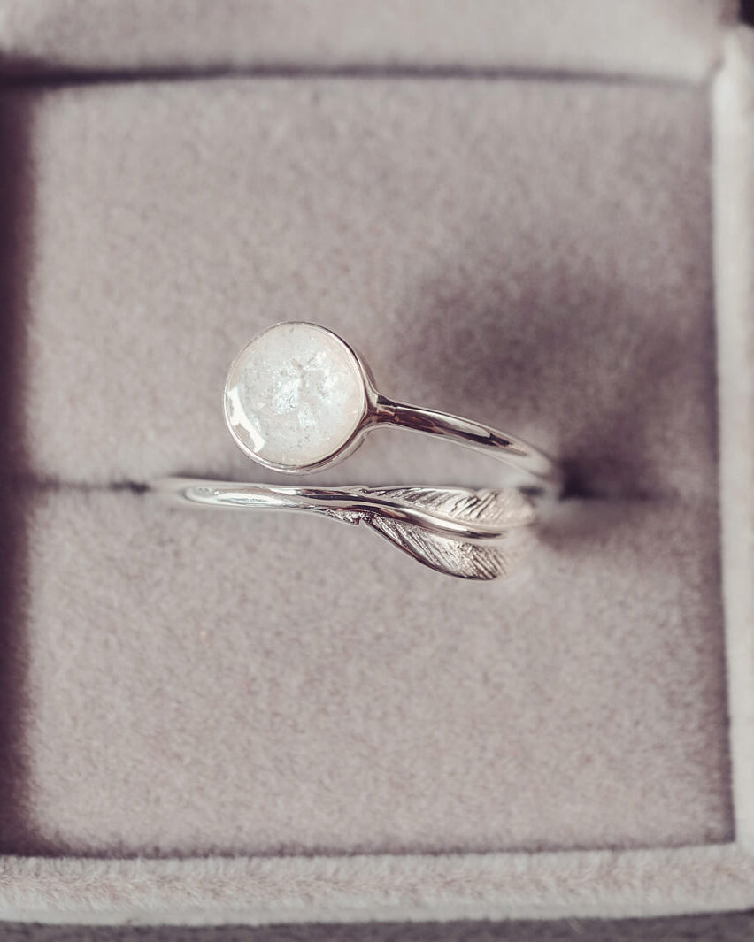 A sterling silver adjustable ring, with a circular bezel filled with clear and white crystals and ashes and a silver feather embellishment