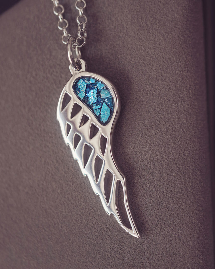 Silver Angel Wing Necklace with blue floating crystals which hold cremation ashes. Shown on a dark background