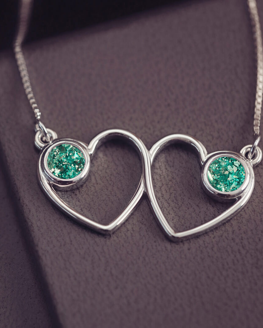 A delicate silver necklace featuring two intertwined heart-shaped pendants, each with a green resin stone which holds crystal and cremation ashes. Displayed against a neutral background.