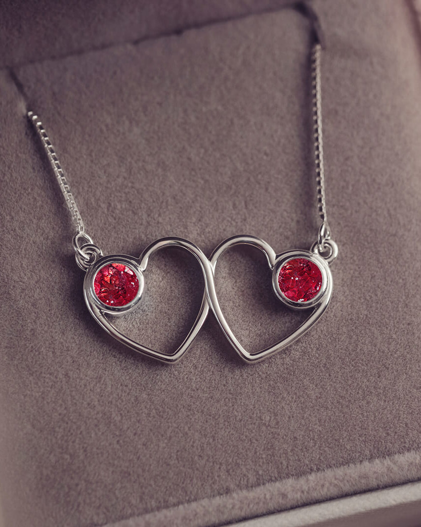 A delicate silver necklace featuring two intertwined heart-shaped pendants, each with a red resin stone which holds crystal and cremation ashes. Displayed against a neutral background.