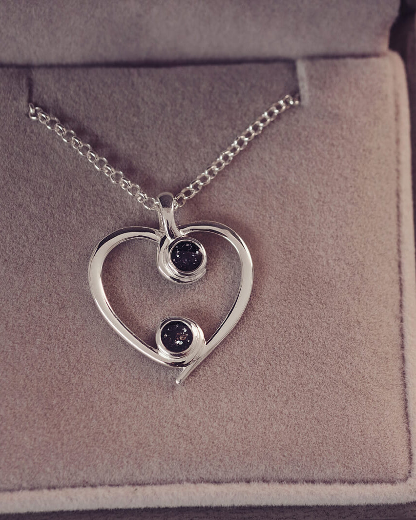 A silver heart-shaped pendant necklace featuring two sparkling black resin stones, holding cremation ashes, displayed against a neutral background.