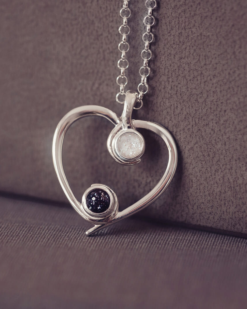 A silver heart-shaped pendant necklace featuring two sparkling black and clear resin stones, holding cremation ashes, displayed against a neutral background.
