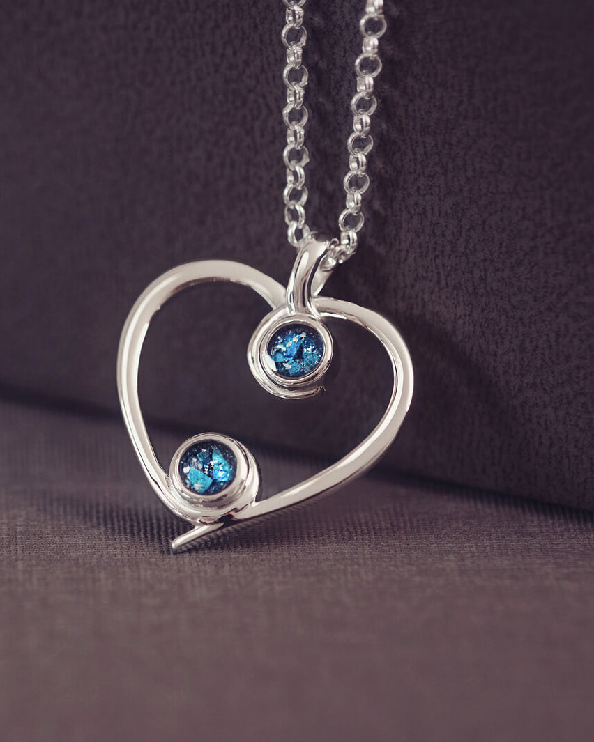 A silver heart-shaped pendant necklace featuring two sparkling blue resin stones, holding cremation ashes, displayed against a neutral background.