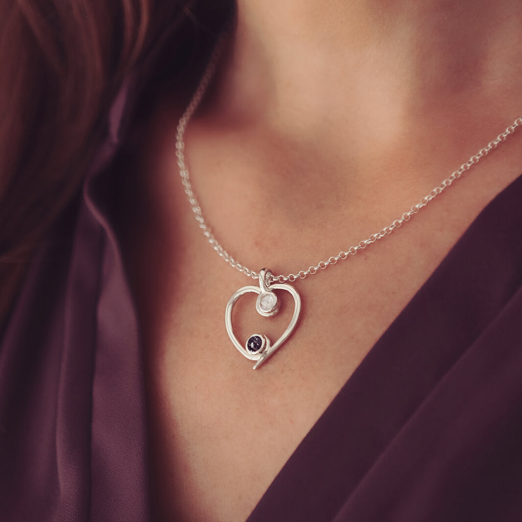 A silver heart-shaped pendant necklace featuring two sparkling resin stones, holding cremation ashes, worn on a ladies neck