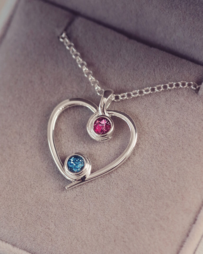 A silver heart-shaped pendant necklace featuring two sparkling pink and blue resin stones, holding cremation ashes, displayed against a neutral background.