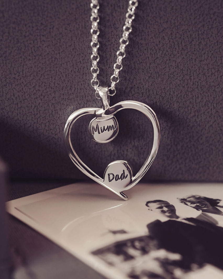 A silver heart-shaped pendant necklace featuring two short names (mum and dad) engraved onto the reverse of each bezel, holding cremation ashes, displayed against a neutral background.