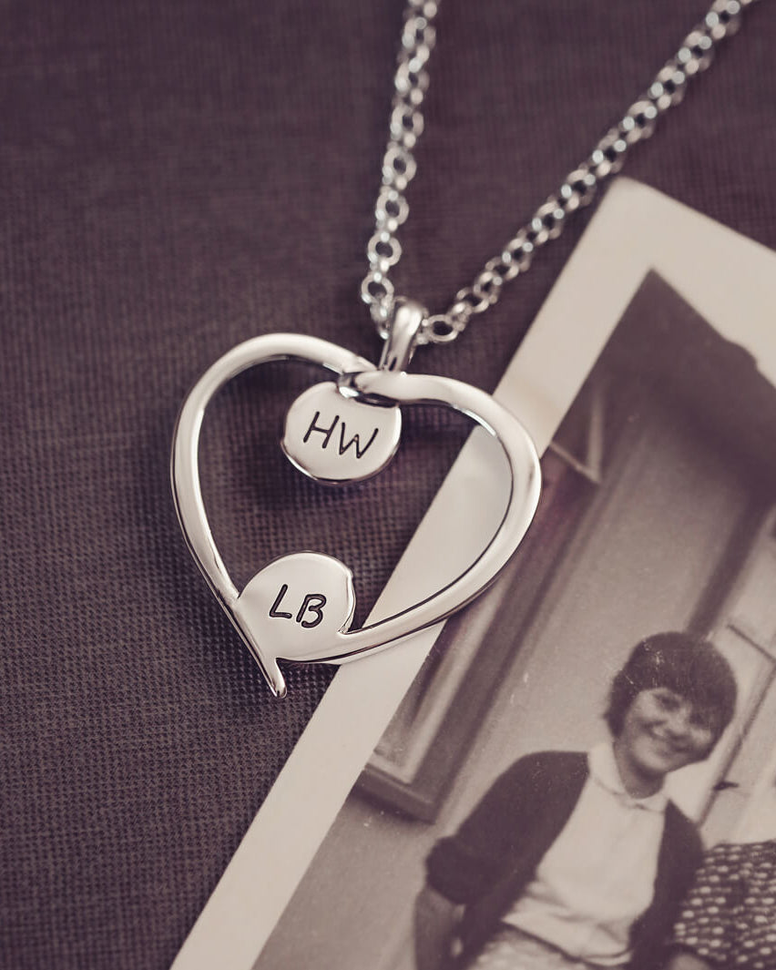 A silver heart-shaped pendant necklace featuring engraved initials on each bezel, holding cremation ashes, displayed against a neutral background.