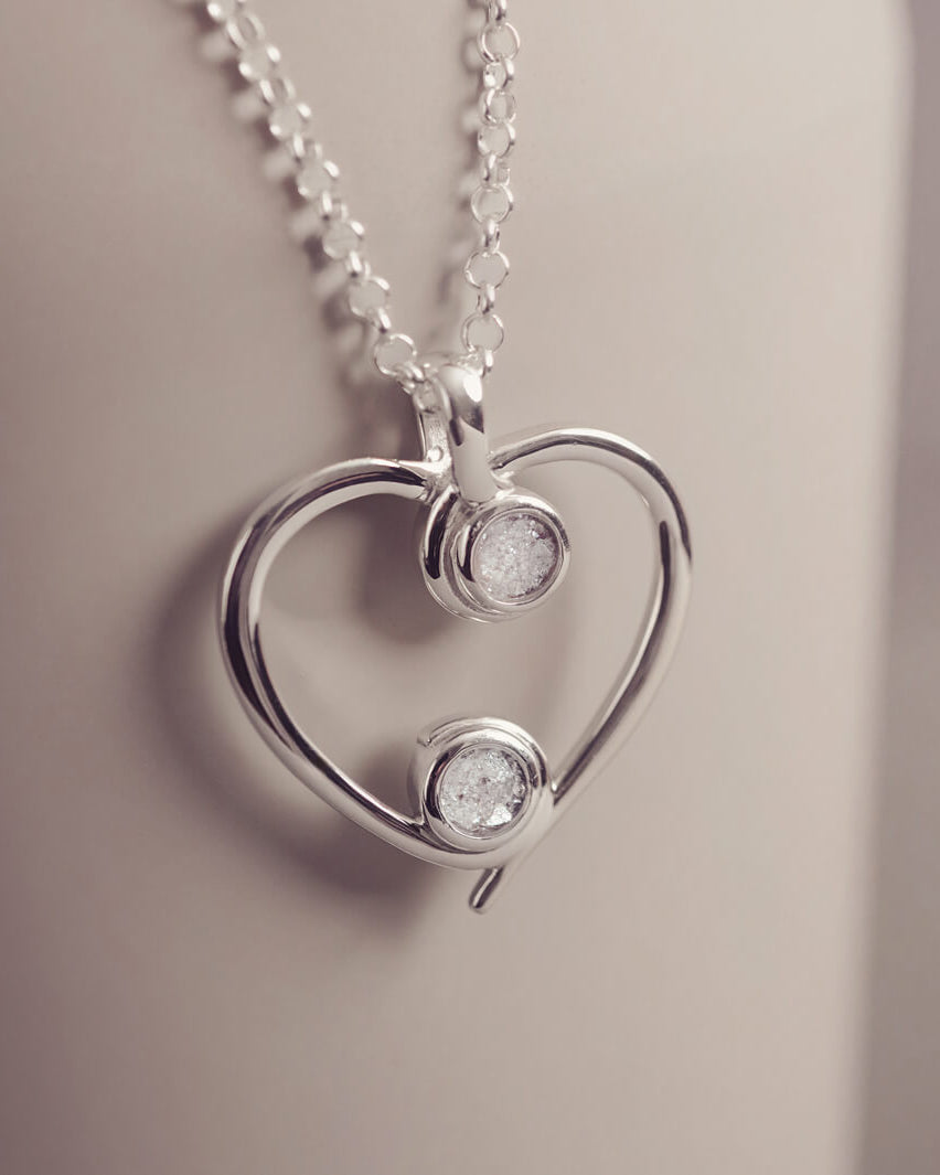 A silver heart-shaped pendant necklace featuring two sparkling clear resin stones, holding cremation ashes, displayed against a neutral background.