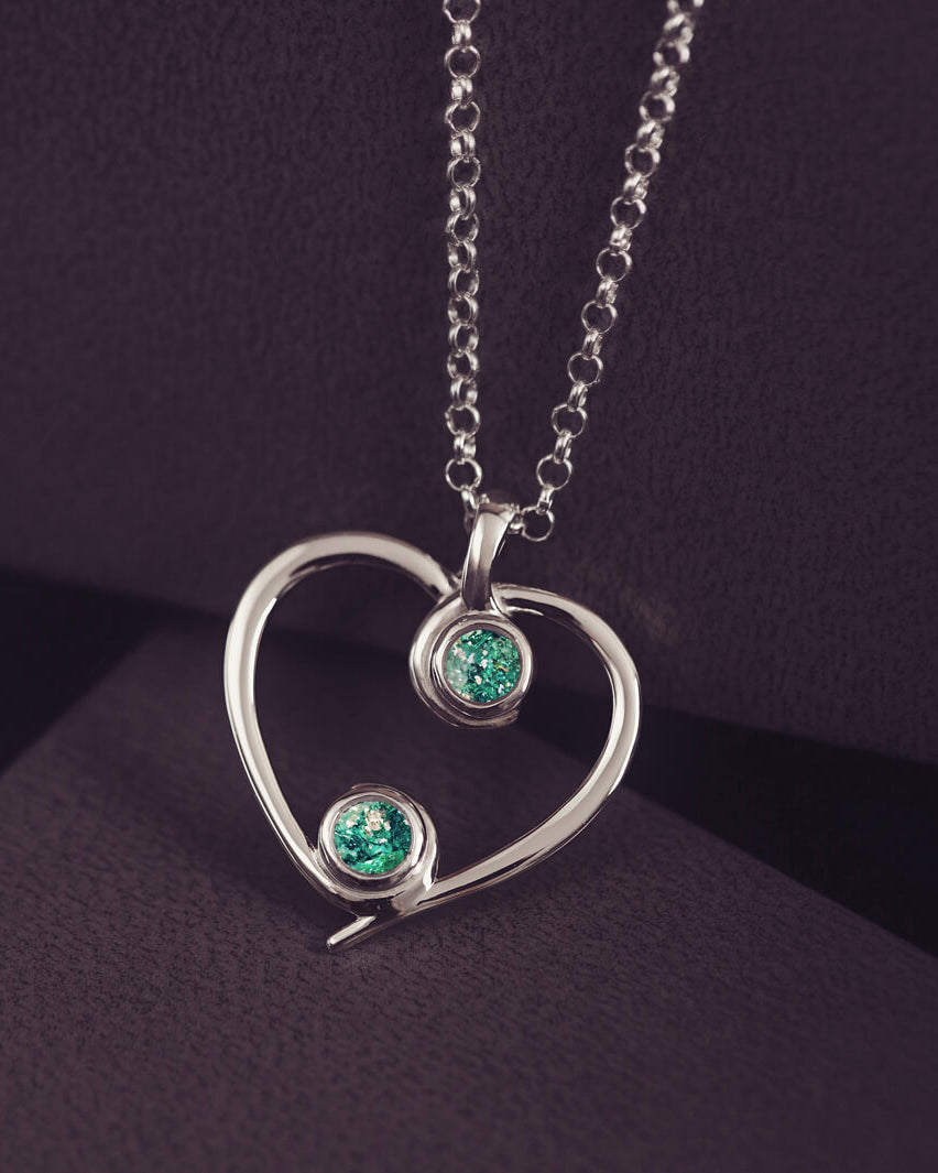 A silver heart-shaped pendant necklace featuring two sparkling green resin stones, holding cremation ashes, displayed against a dark background.