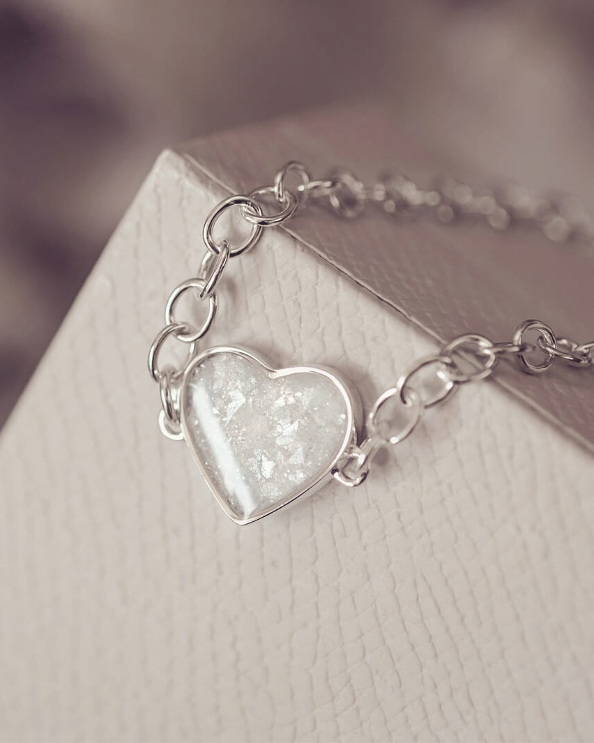 silver bracelet & heart charm, holding clear crystal, resin & cremation ashes. laying on a grey jewellery box