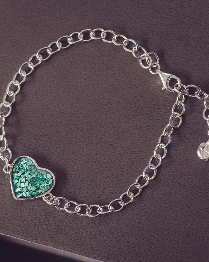 silver bracelet & heart charm, holding green crystal, resin & cremation ashes. laying on a black back ground