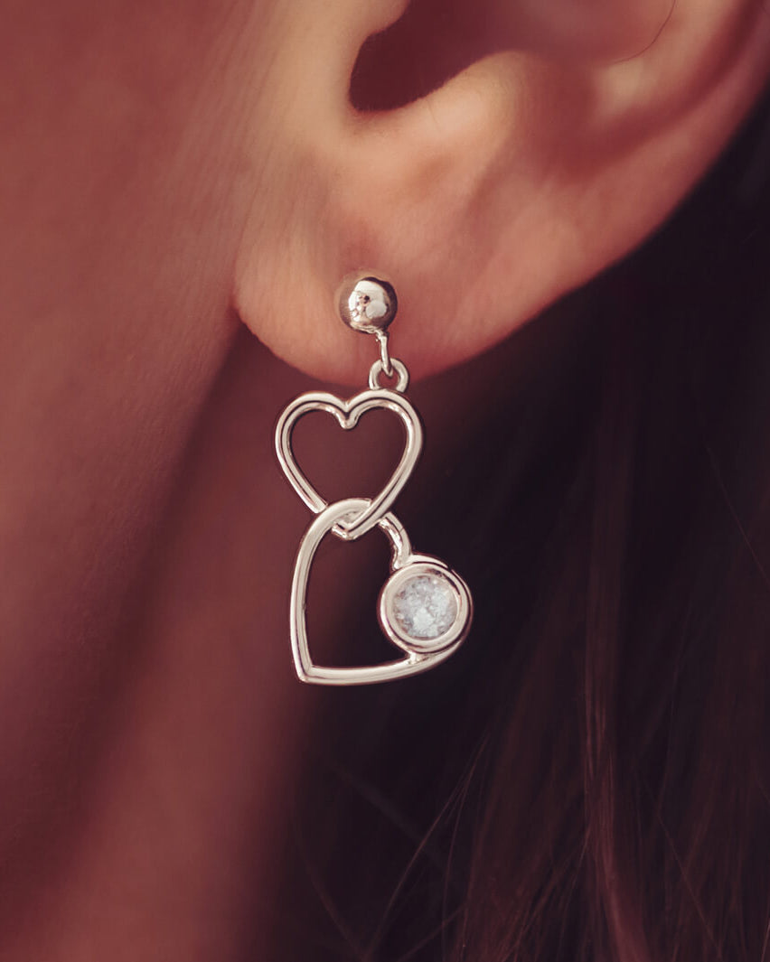 Silver Heart Dangle Earrings, holding cremation ashes, with clear crystals. Worn in a ladies ear.