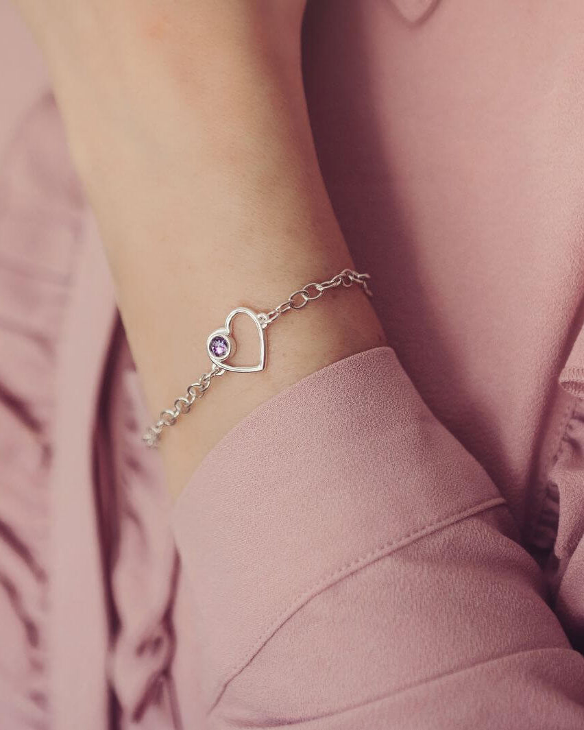 Silver bracelet with a purple heart cremation ashes charm on a wrist against a pink background