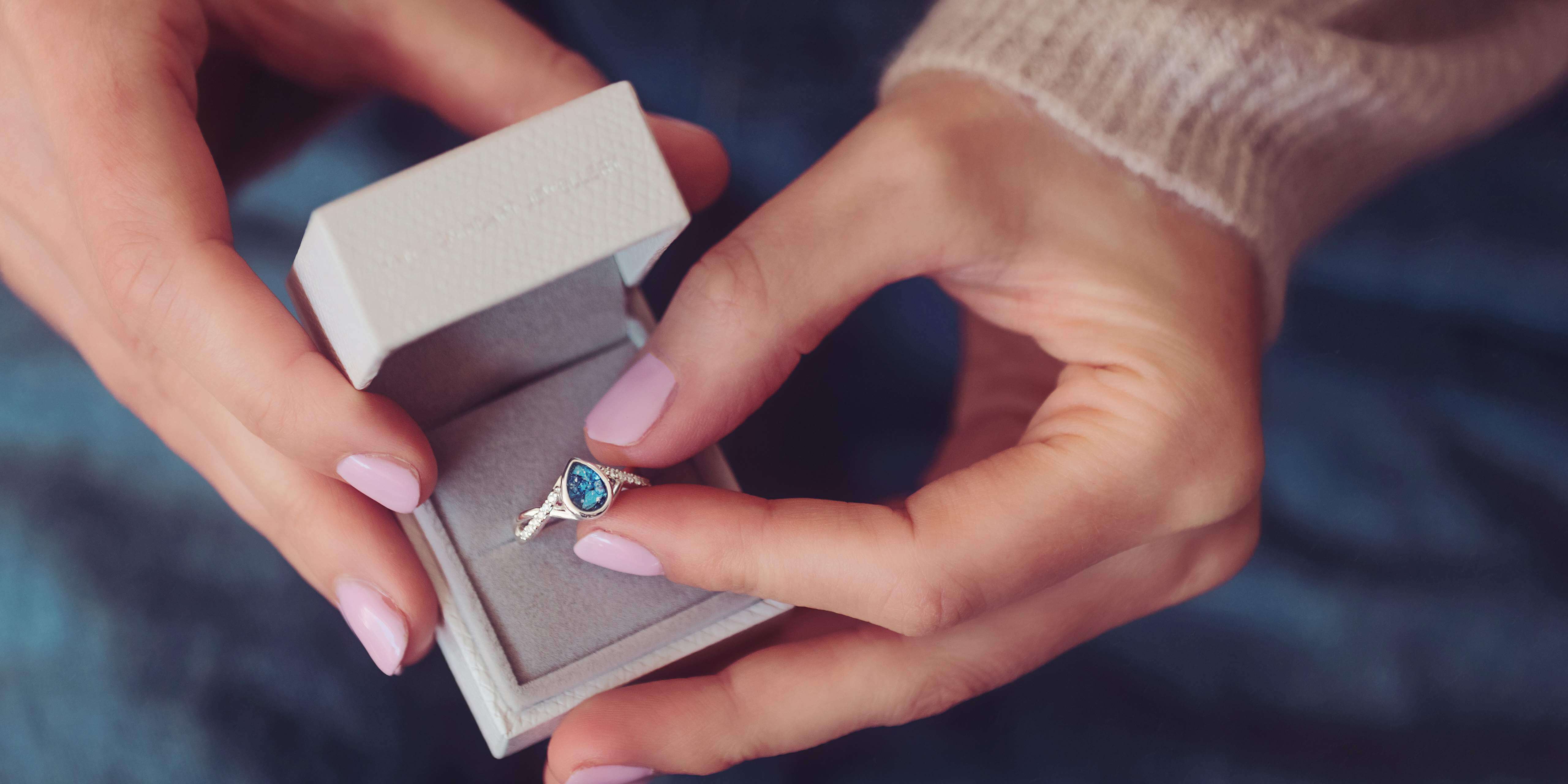 A blue teardrop ring in a grey box being held in two female hands