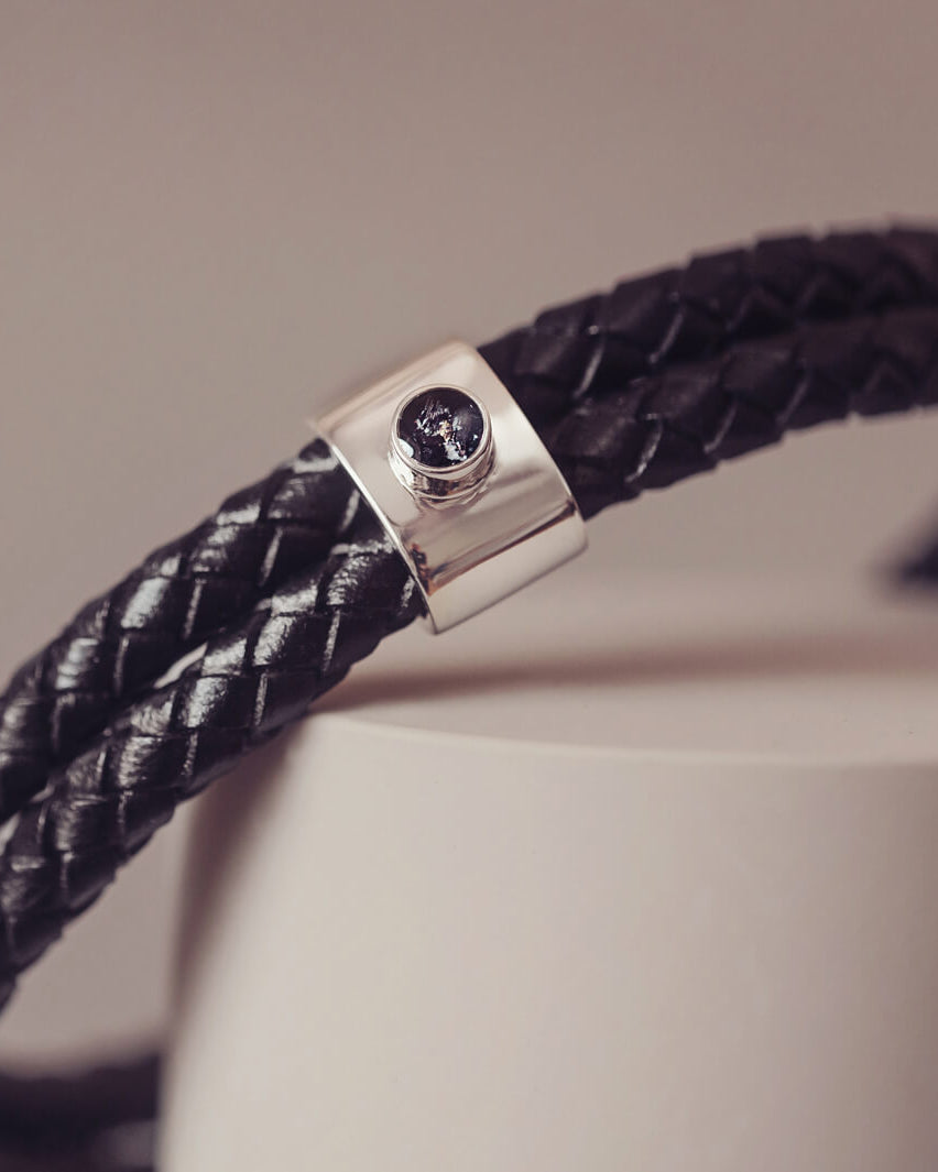 Black braided leather bracelet featuring a silver charm with a small black memorial stone set in the centre, displayed on a grey surface.