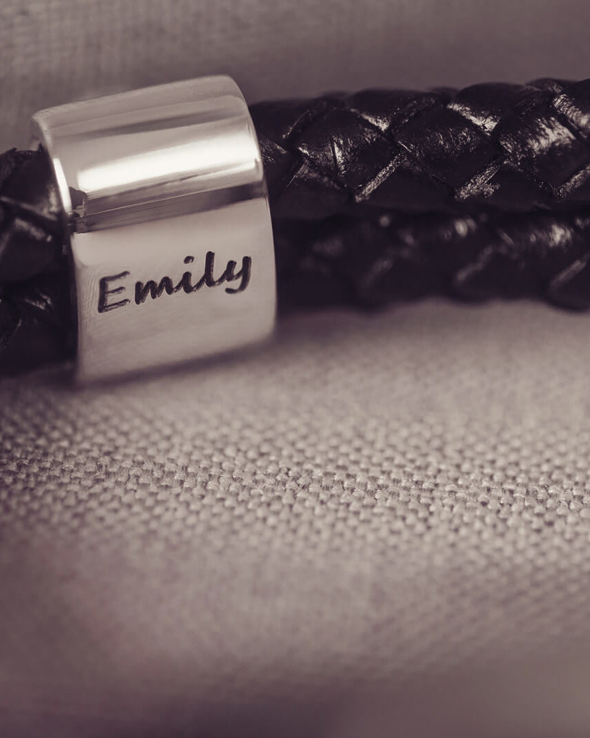 Black braided leather bracelet featuring a silver charm with the name 'Emily' engraved on it in black, displayed on a grey surface.