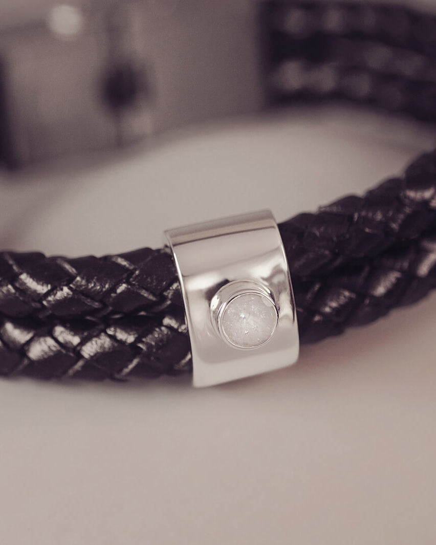Black braided leather bracelet featuring a silver charm with a small clear memorial stone set in the centre, displayed on a grey surface.