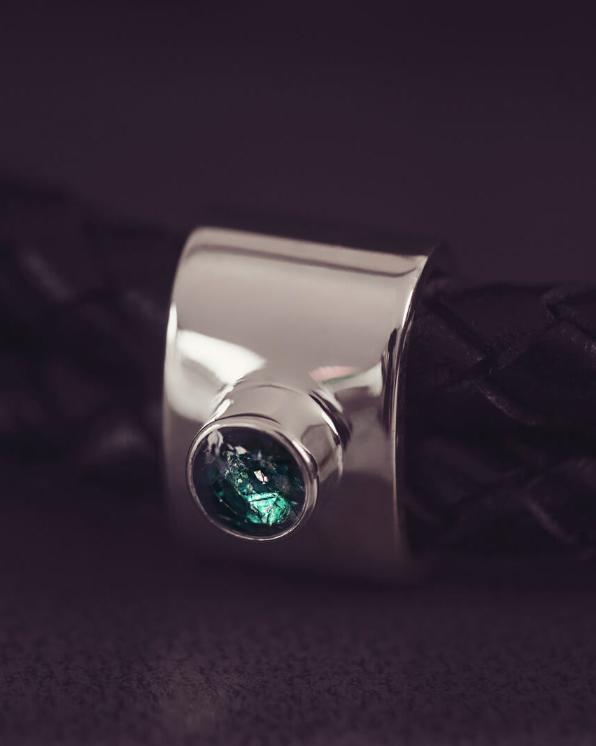 Black braided leather bracelet featuring a silver charm with a small green memorial stone set in the centre, displayed on a grey surface.