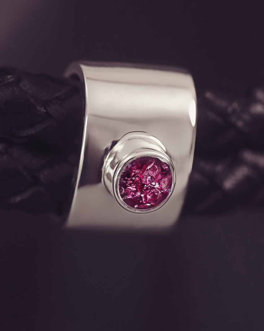 Black braided leather bracelet featuring a silver charm with a small pink memorial stone set in the centre, displayed on a grey surface.