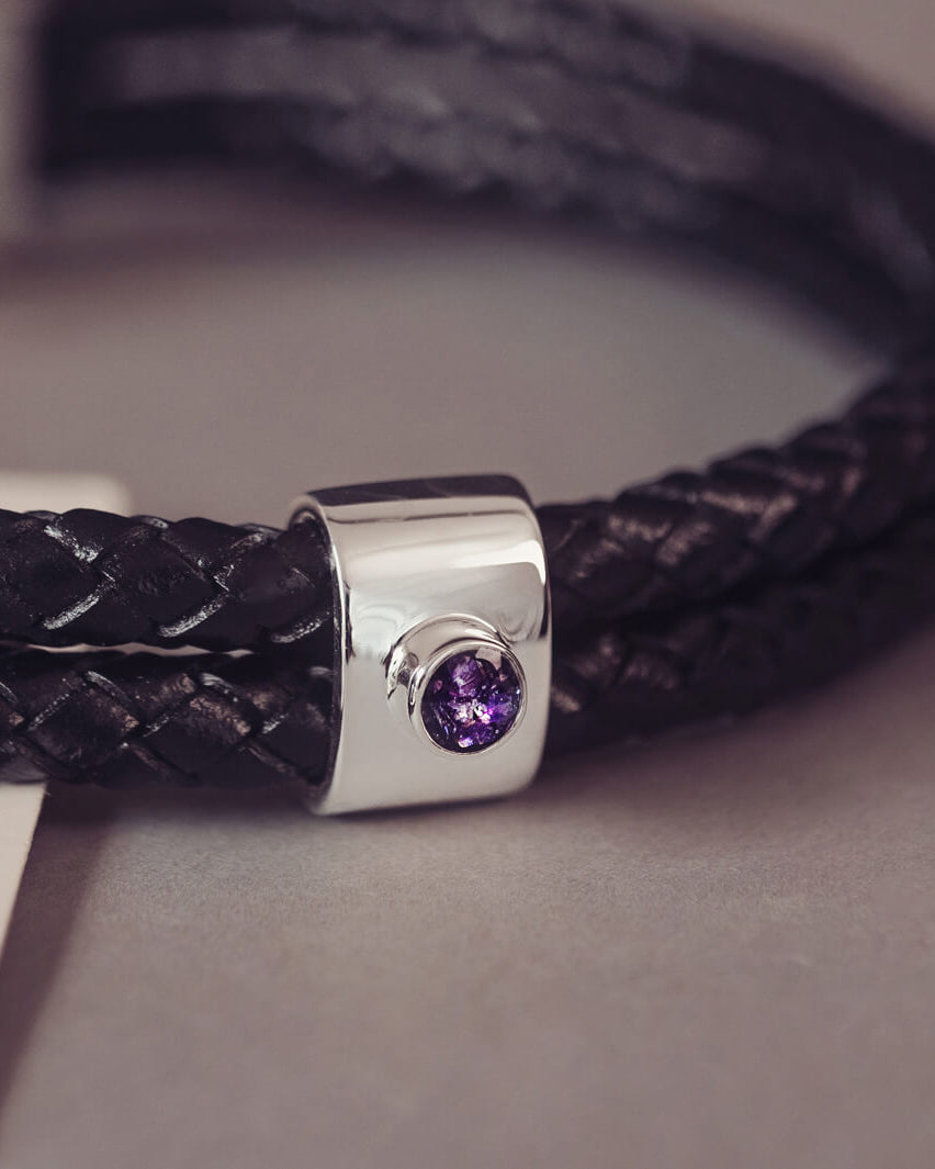 Black braided leather bracelet featuring a silver charm with a small purple memorial stone set in the centre, displayed on a grey surface.