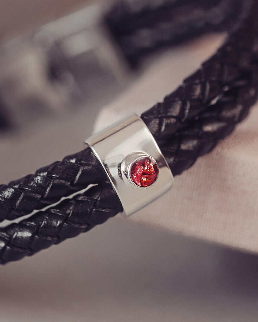Black braided leather bracelet featuring a silver charm with a small red memorial stone set in the center, displayed on a grey surface.