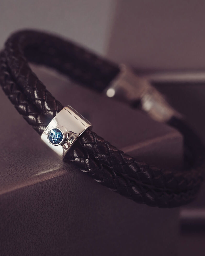 Black braided leather bracelet featuring a silver charm with a small blue memorial stone set in the centre, displayed on a grey surface.