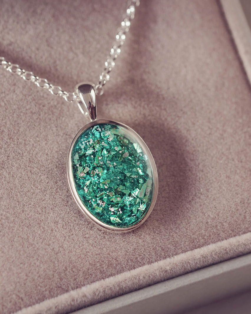 Sterling silver oval necklace with a central section filled with memorial ashes and green crushed crystals, displayed on a grey fabric background