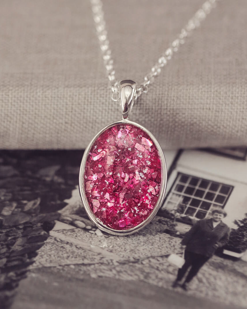 Sterling silver oval necklace with a central section filled with memorial ashes and pink crushed crystals, displayed on a background of a black and white photo, showing a lady stood in front of a house