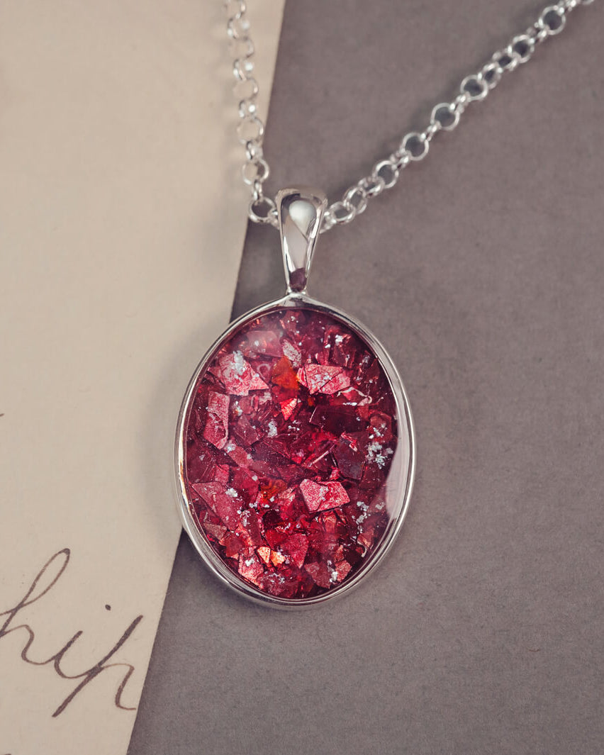 An oval shaped sterling silver pendant, on a silver belcher chain, filed with red crushed crystal and cremation ashes