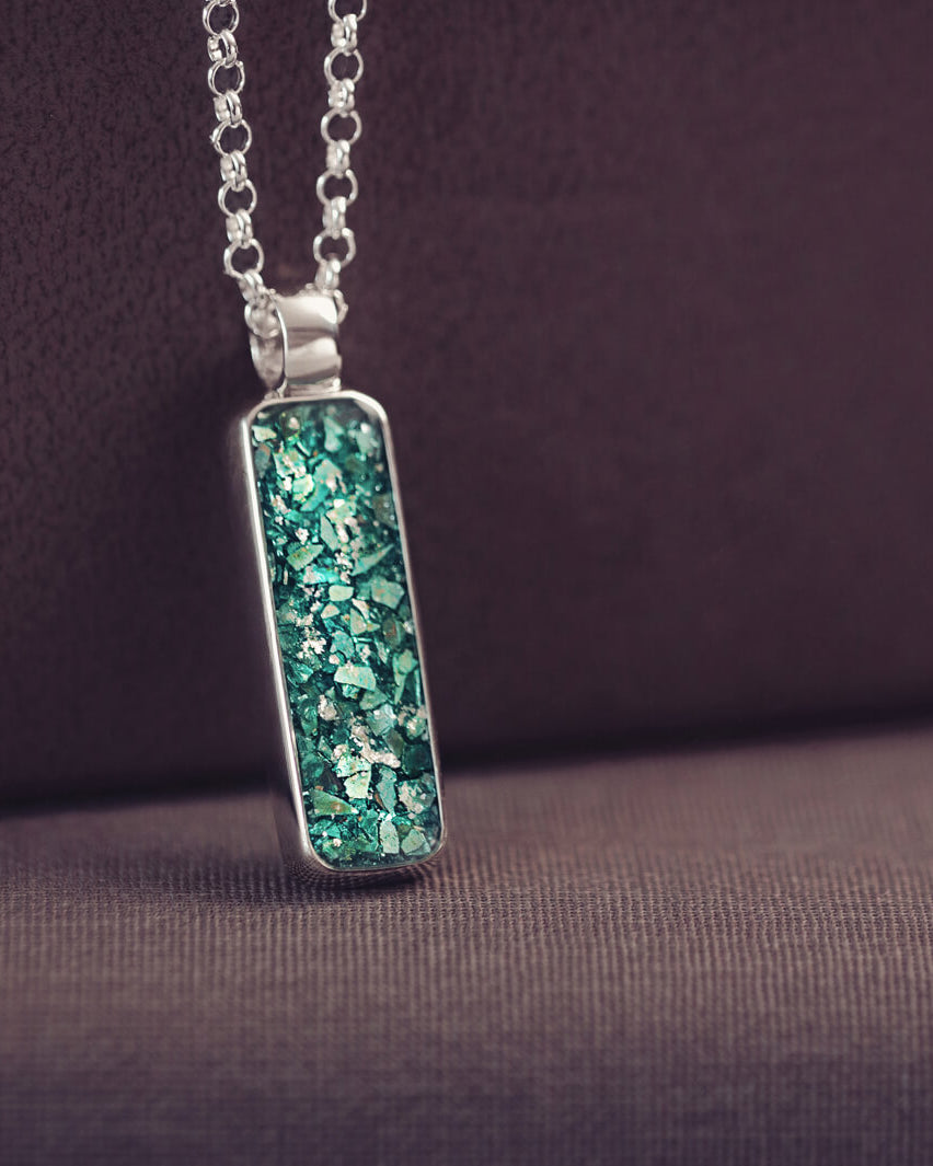 A sophisticated silver necklace featuring a rectangular pendant filled with shimmering green crystal and resin, delicately layered with cremation ashes, showcased against a dark background, on a silver belcher chain.