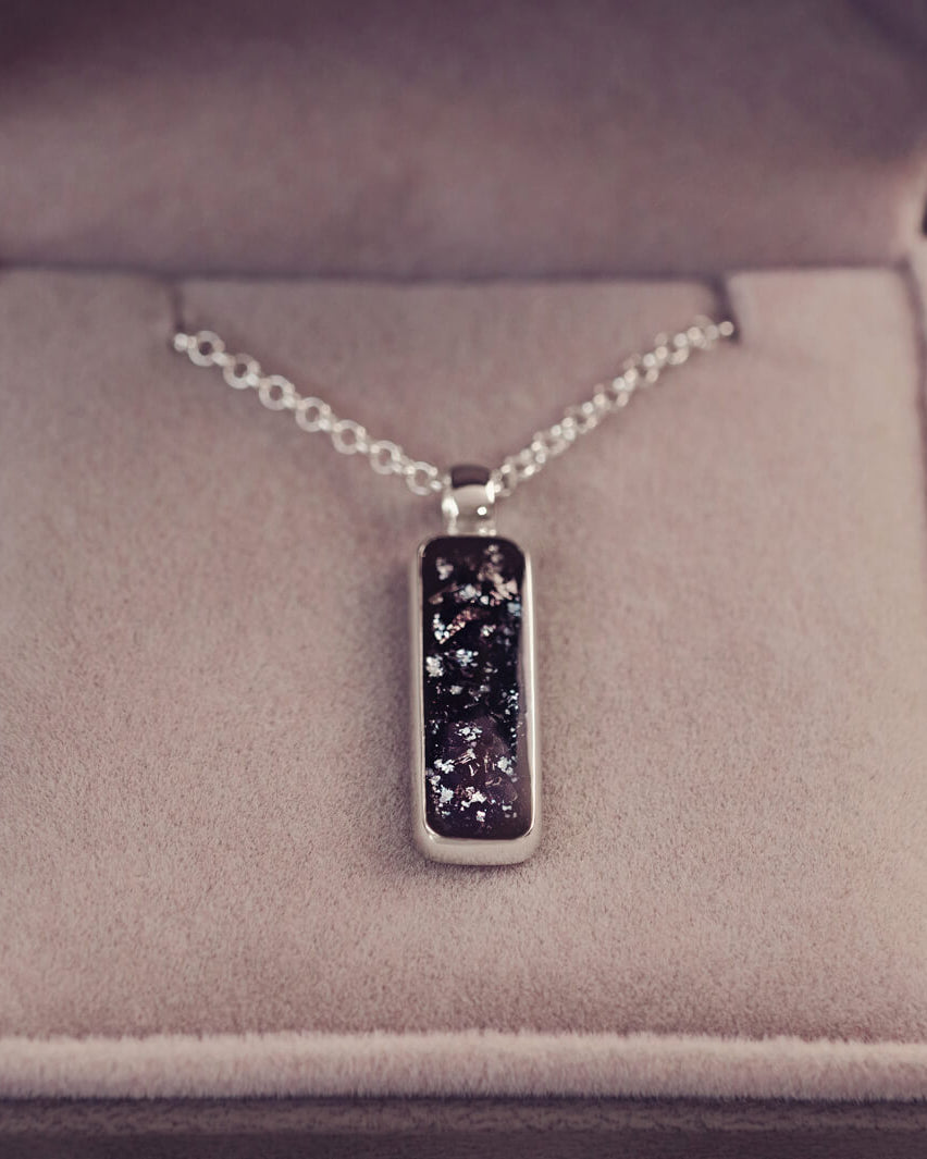 A sophisticated silver necklace featuring a rectangular pendant filled with shimmering black crystal and resin, delicately layered with cremation ashes, showcased against a light background, on a silver belcher chain.