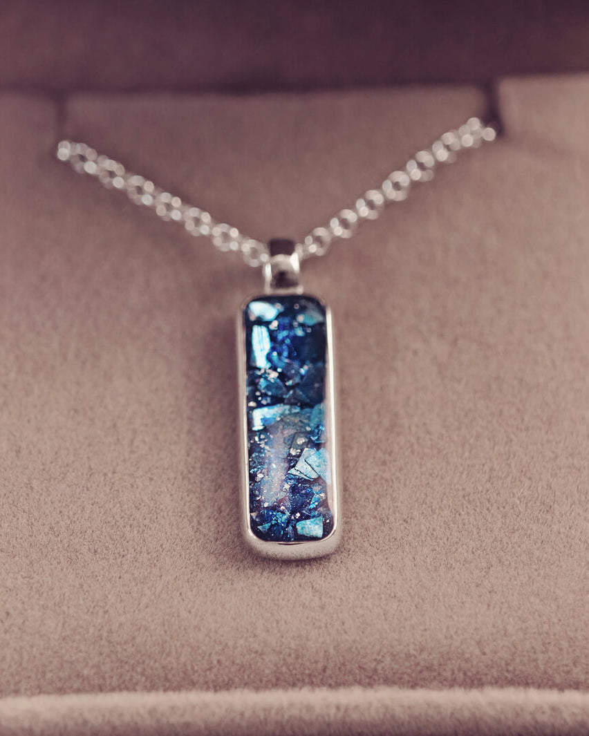 A sophisticated silver necklace featuring a rectangular pendant filled with shimmering blue crystal and resin, delicately layered with cremation ashes, showcased against a light background, on a silver belcher chain.