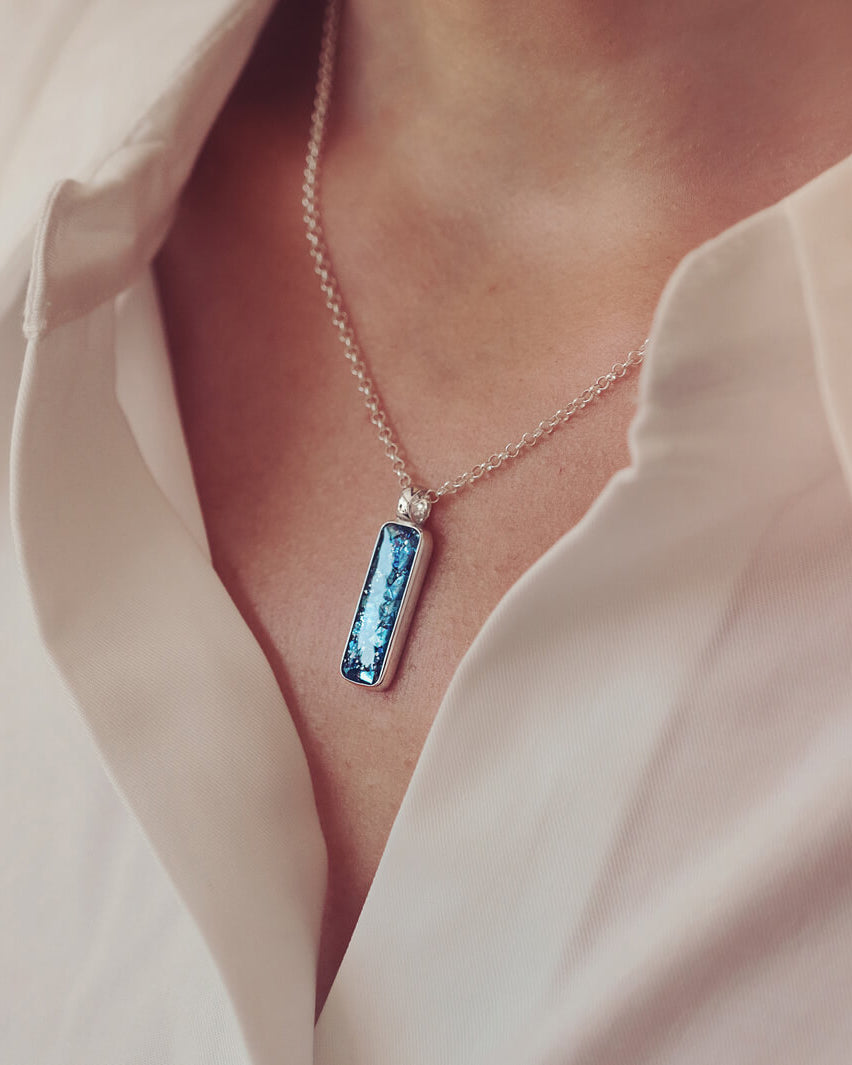 A sterling silver rectangle necklace, holding blue crystal and cremation ashes, on a silver chain being worn around a ladies neck