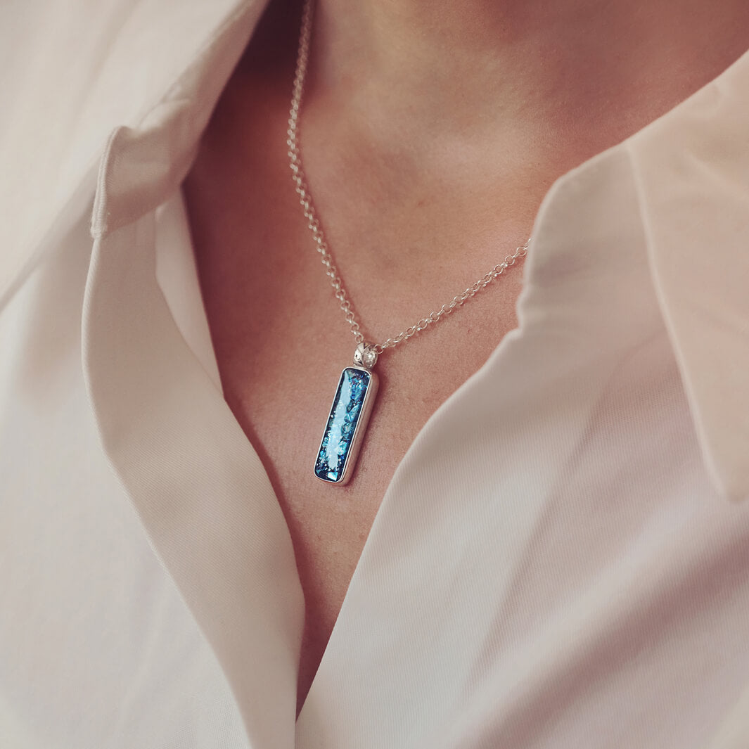A sterling silver rectangle necklace, holding blue crystal and cremation ashes, on a silver chain being worn around a ladies neck
