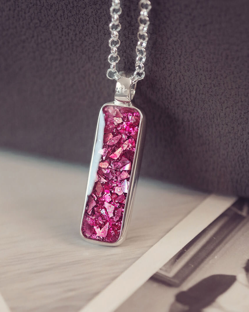 A sophisticated silver necklace featuring a rectangular pendant filled with shimmering pink crystal and resin, delicately layered with cremation ashes, showcased against a dark background, on a silver belcher chain.