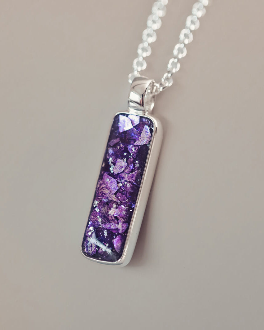 A sophisticated silver necklace featuring a rectangular pendant filled with shimmering purple crystal and resin, delicately layered with cremation ashes, showcased against a light background, on a silver belcher chain.