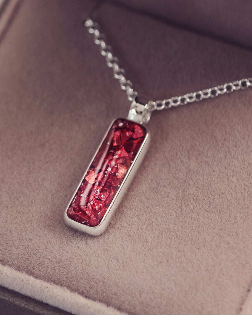 A sophisticated silver necklace featuring a rectangular pendant filled with shimmering red crystal and resin, delicately layered with cremation ashes, showcased against a dark background, on a silver belcher chain.
