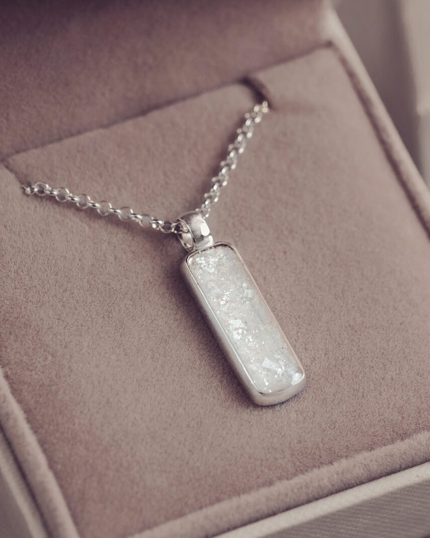 A sophisticated silver necklace featuring a rectangular pendant filled with shimmering white resin, delicately layered with cremation ashes, showcased against a light background.