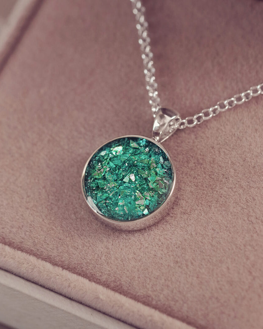 A round sterling silver necklace charm, hanging on a sterling silver chain. The charm is filled with green crystal and cremation ashes