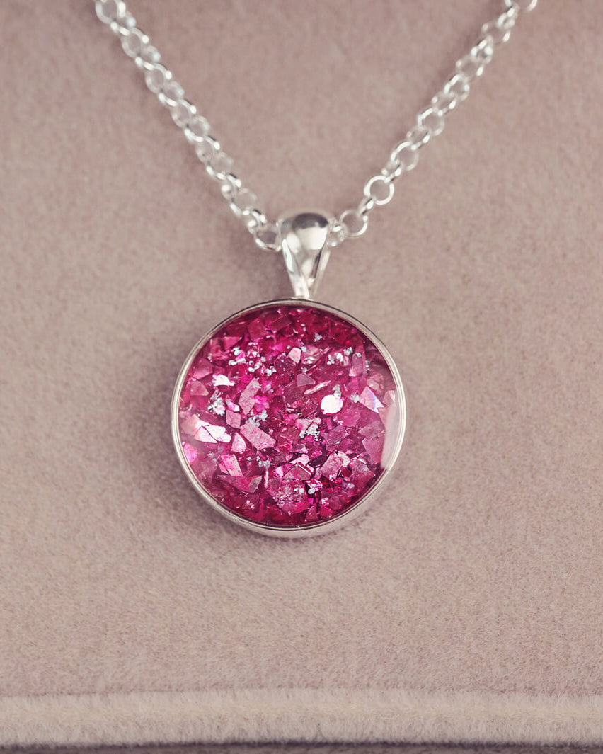 A round sterling silver necklace charm, hanging on a sterling silver chain. The charm is filled with pink crystal and cremation ashes