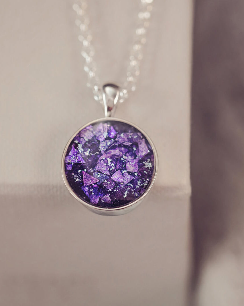 A round sterling silver pendant, hanging from a silver belcher chain, filled with purple crushed crystal and cremation ashes.