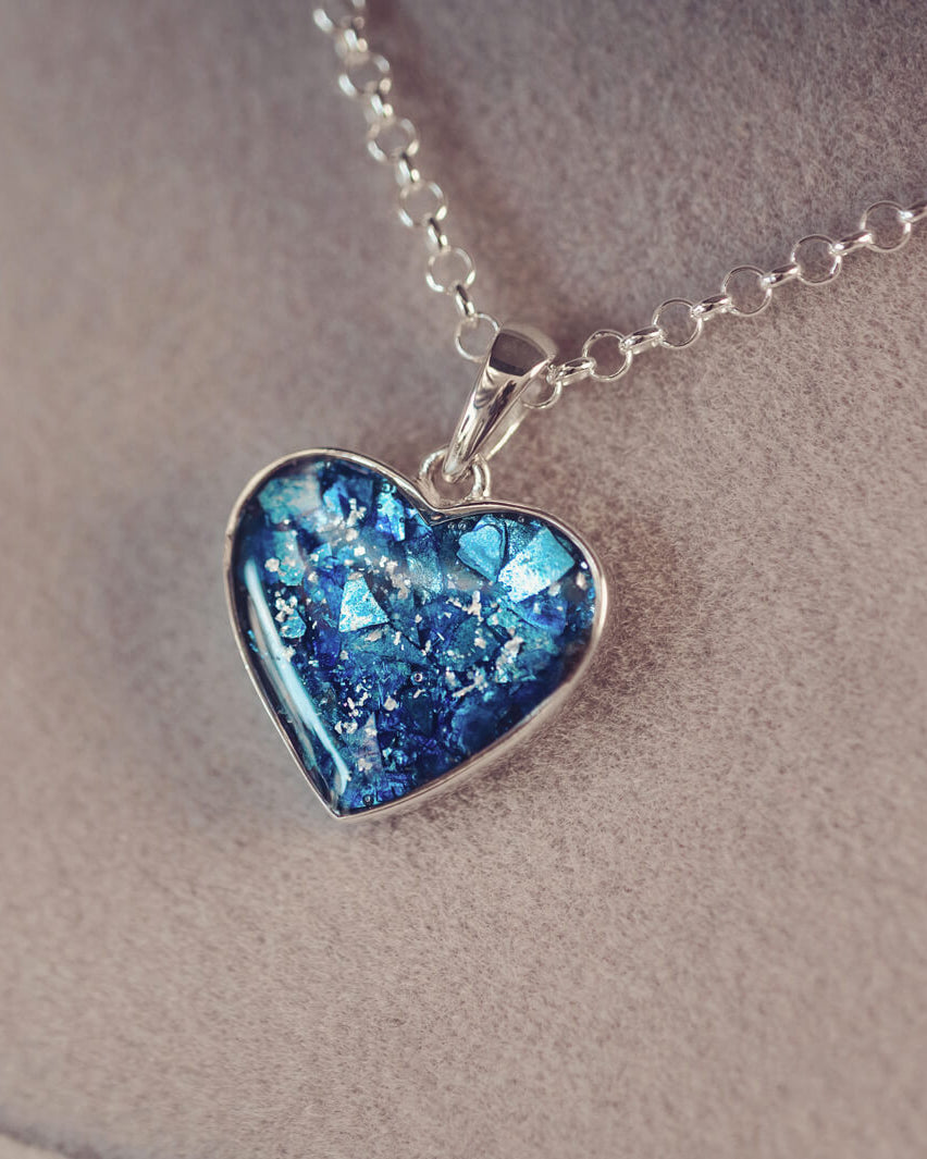 Sterling silver heart necklace with a central section filled with memorial ashes and blue crushed crystals, displayed on a grey fabric background.
