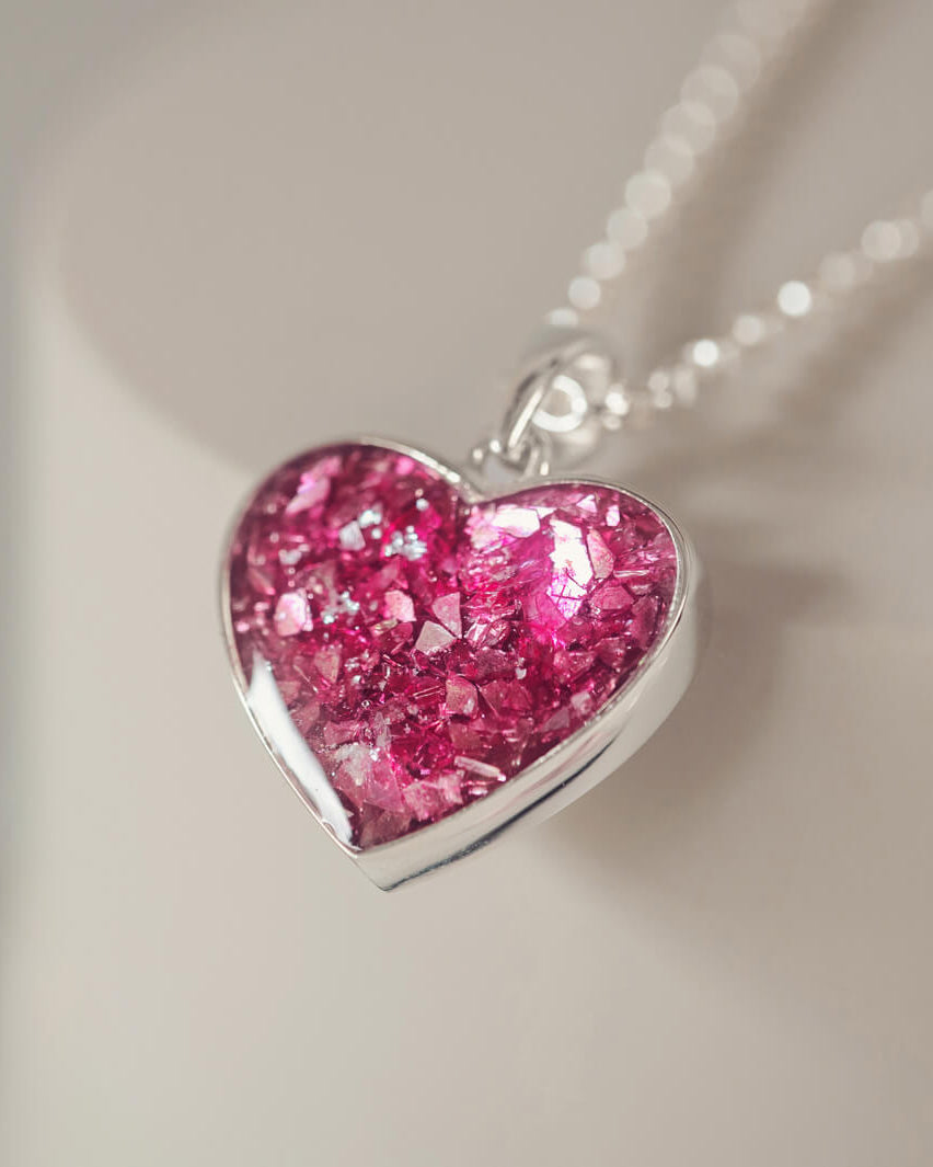 Sterling silver heart necklace with a central section filled with memorial ashes and pink crushed crystals, displayed on a blurred white background
