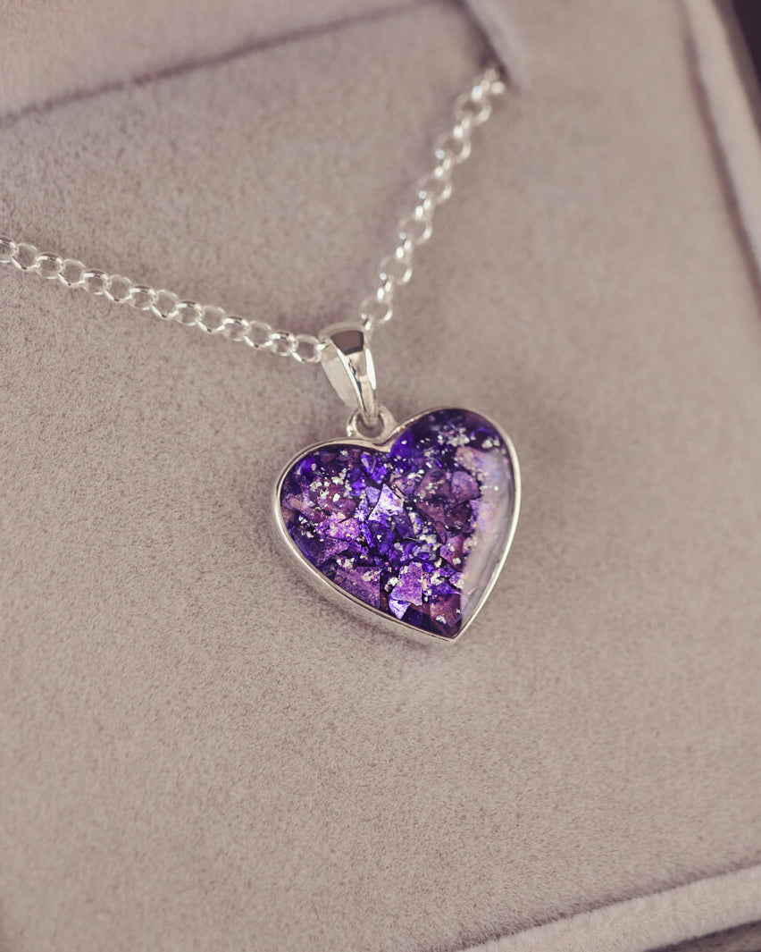 Sterling silver heart necklace with a central section filled with memorial ashes and purple crushed crystals, displayed on a grey fabric background.