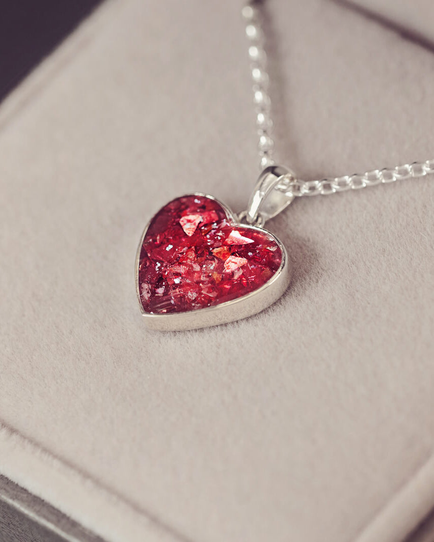 Sterling silver heart necklace with a central section filled with memorial ashes and red crushed crystals, displayed on a grey fabric background.