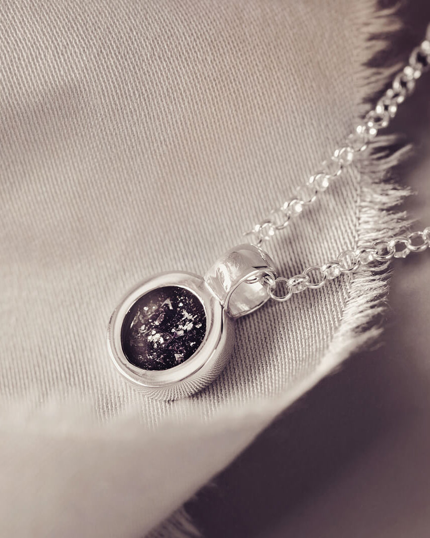 Sterling silver round necklace with a central section filled with memorial ashes and black crushed crystals, displayed on a grey fabric